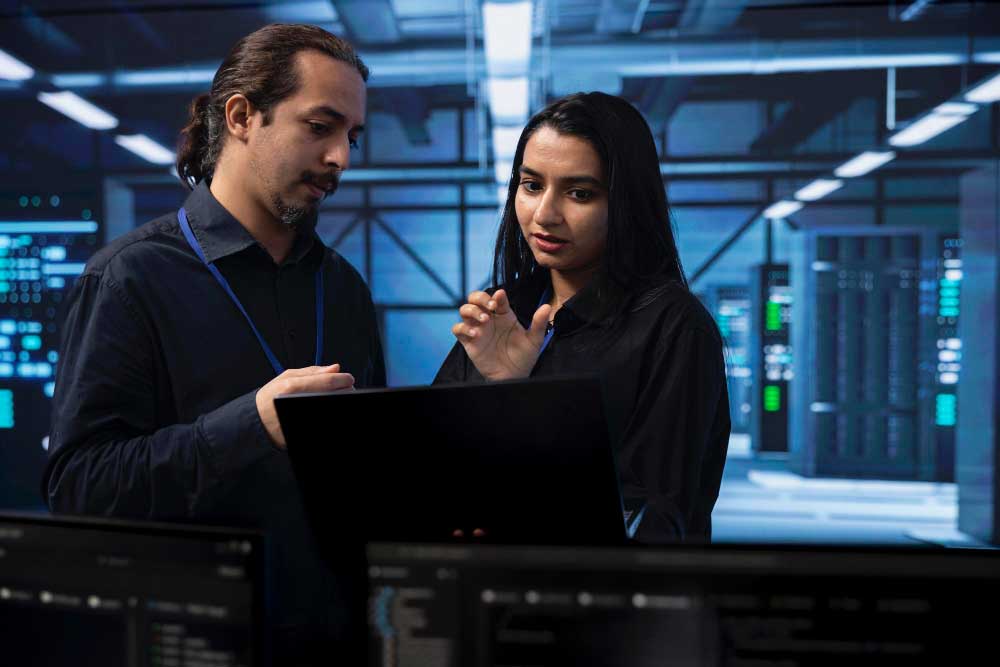 Zwei IT-Spezialisten besprechen Daten auf einem Laptop in einem Serverraum mit leuchtenden Bildschirmen.