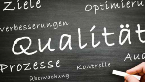 Hand schreibt Wörter wie Qualität, Ziele und Prozesse mit Kreide auf eine Tafel.