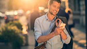 Lächelnder Mann, der auf seinem Smartphone in der sonnigen Innenstadt tippt und eine Jacke über die Schulter trägt.