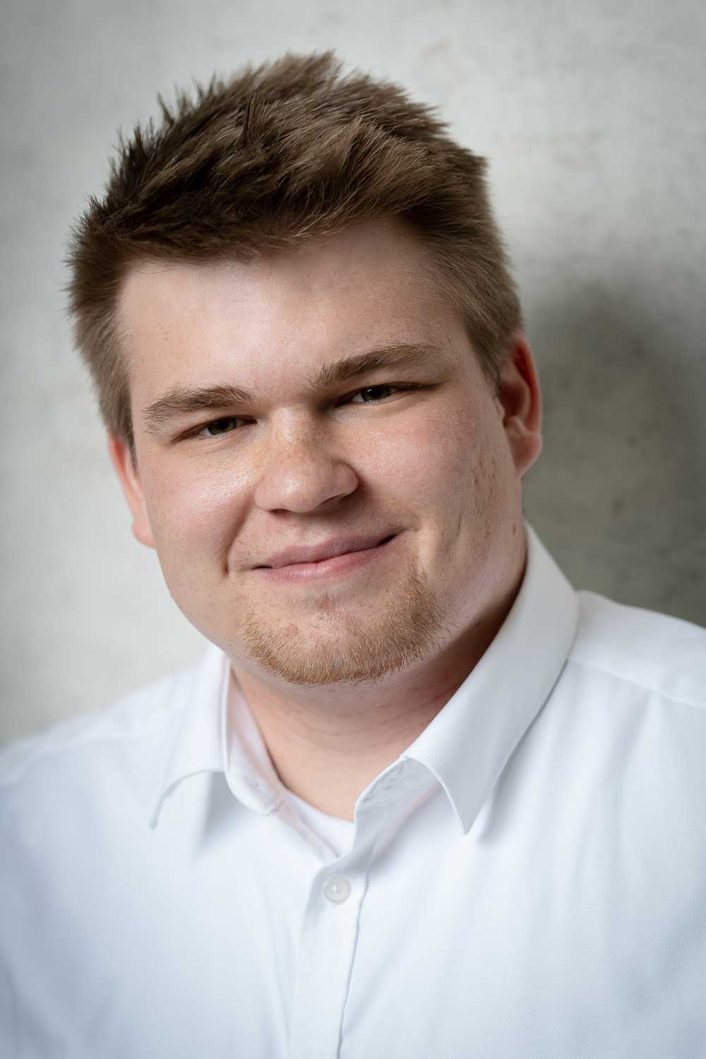 Junger Mann mit kurzem braunem Haar und leichtem Bart lächelt und trägt ein weißes Hemd.