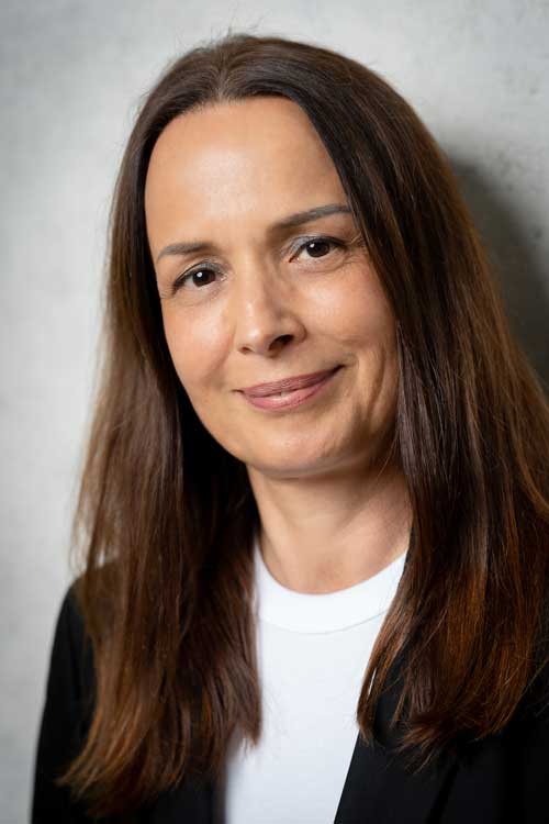 Nahaufnahme einer lächelnden Frau mit langen, braunen Haaren vor grauem Hintergrund.