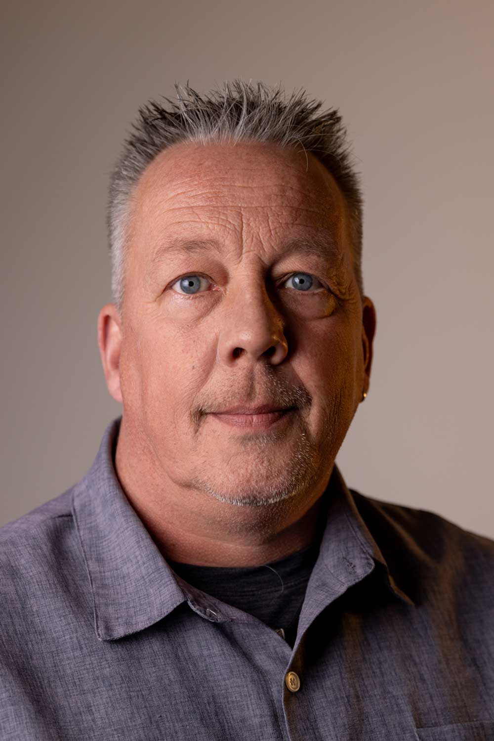 Porträt eines älteren Mannes mit kurz geschnittenem grauem Haar und blauem Hemd vor neutralem Hintergrund.