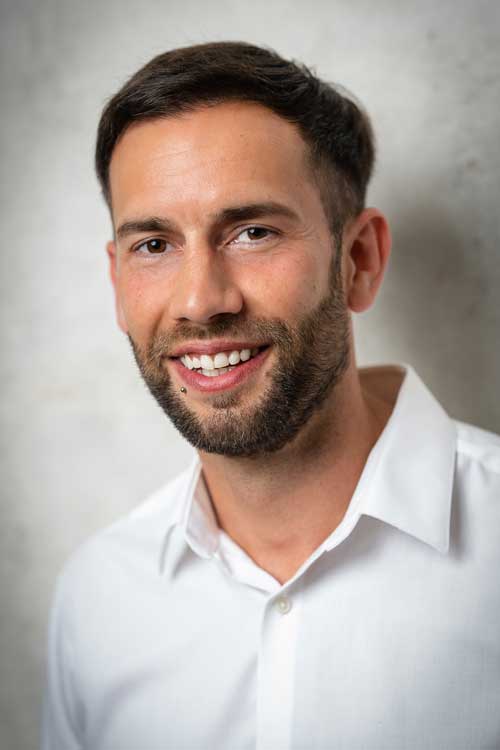 Junger Mann mit kurzem braunem Haar, Bart und Lippenpiercing lächelt, trägt ein weißes Hemd vor grauem Hintergrund.