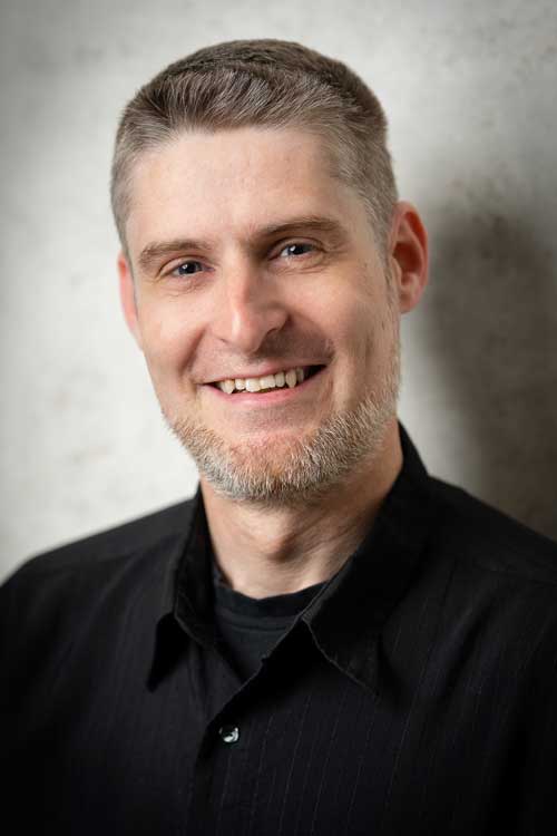 Porträt eines lächelnden Mannes mit kurzem grauem Haar und Bart vor einem hellen Hintergrund, er trägt ein schwarzes Hemd.