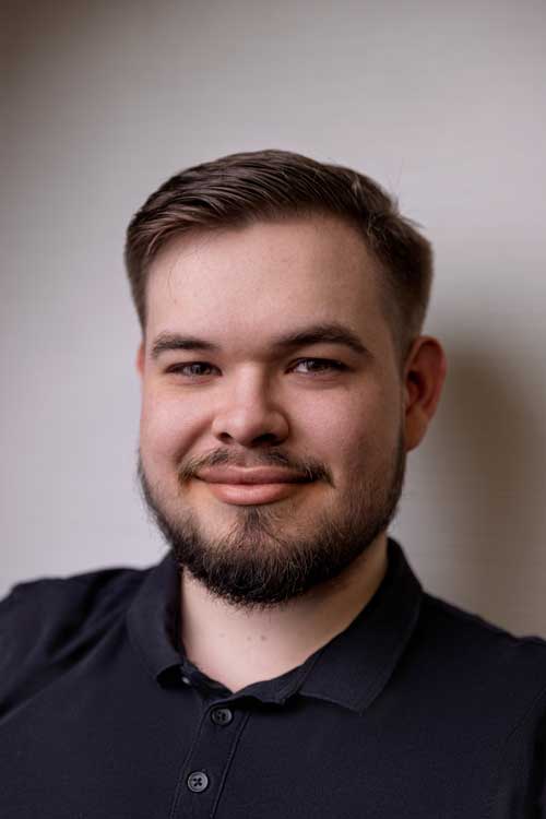 Porträt eines lächelnden bärtigen Mannes mit kurzem braunem Haar und schwarzem Polohemd vor grauem Hintergrund.