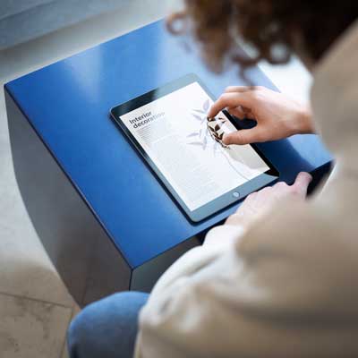Person liest Artikel über Internetdekoration auf einem Tablet, der auf einem blauen Tisch liegt.