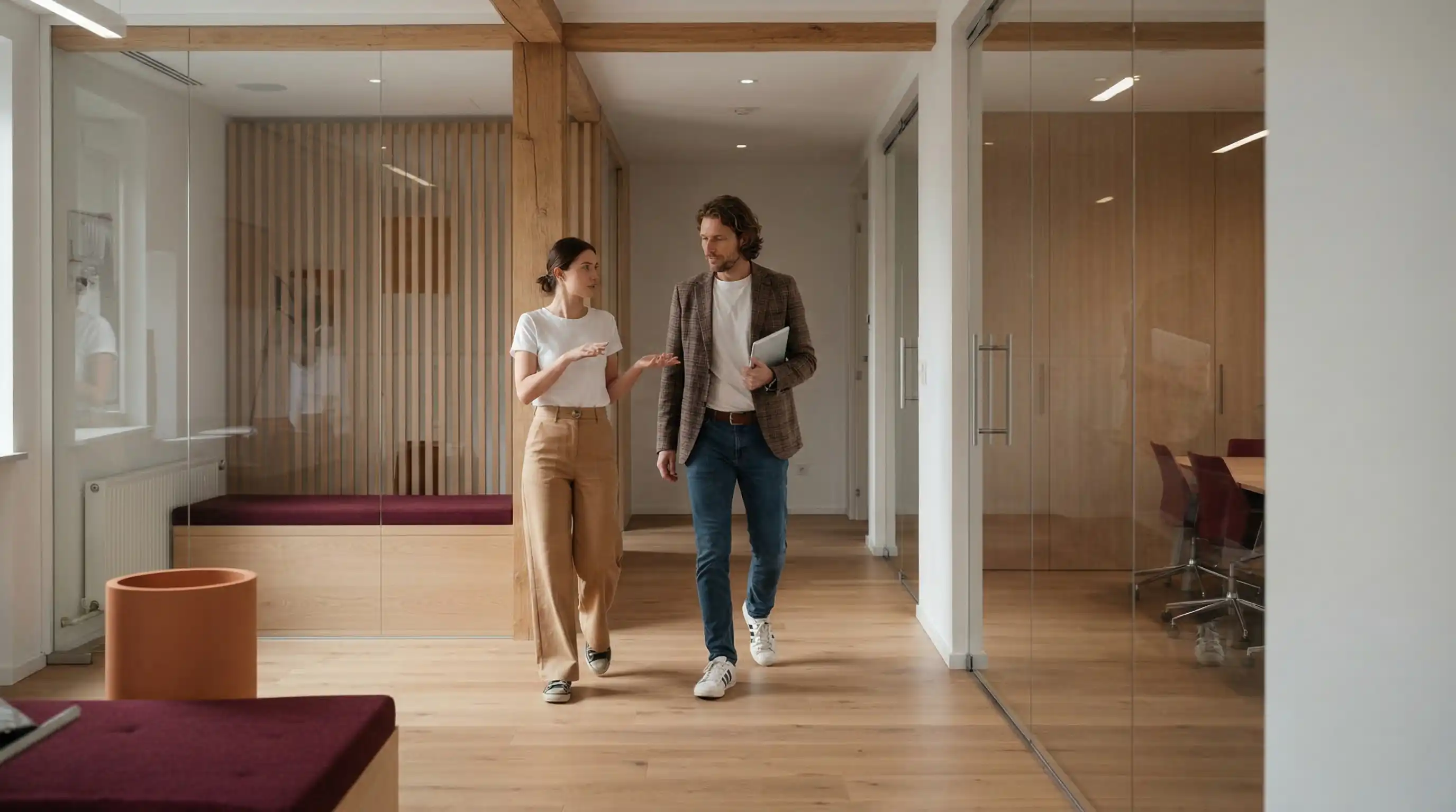 Zwei Personen gehen in einem modernen Büroflur mit Holzfußboden und Glaswänden, während sie sprechen.