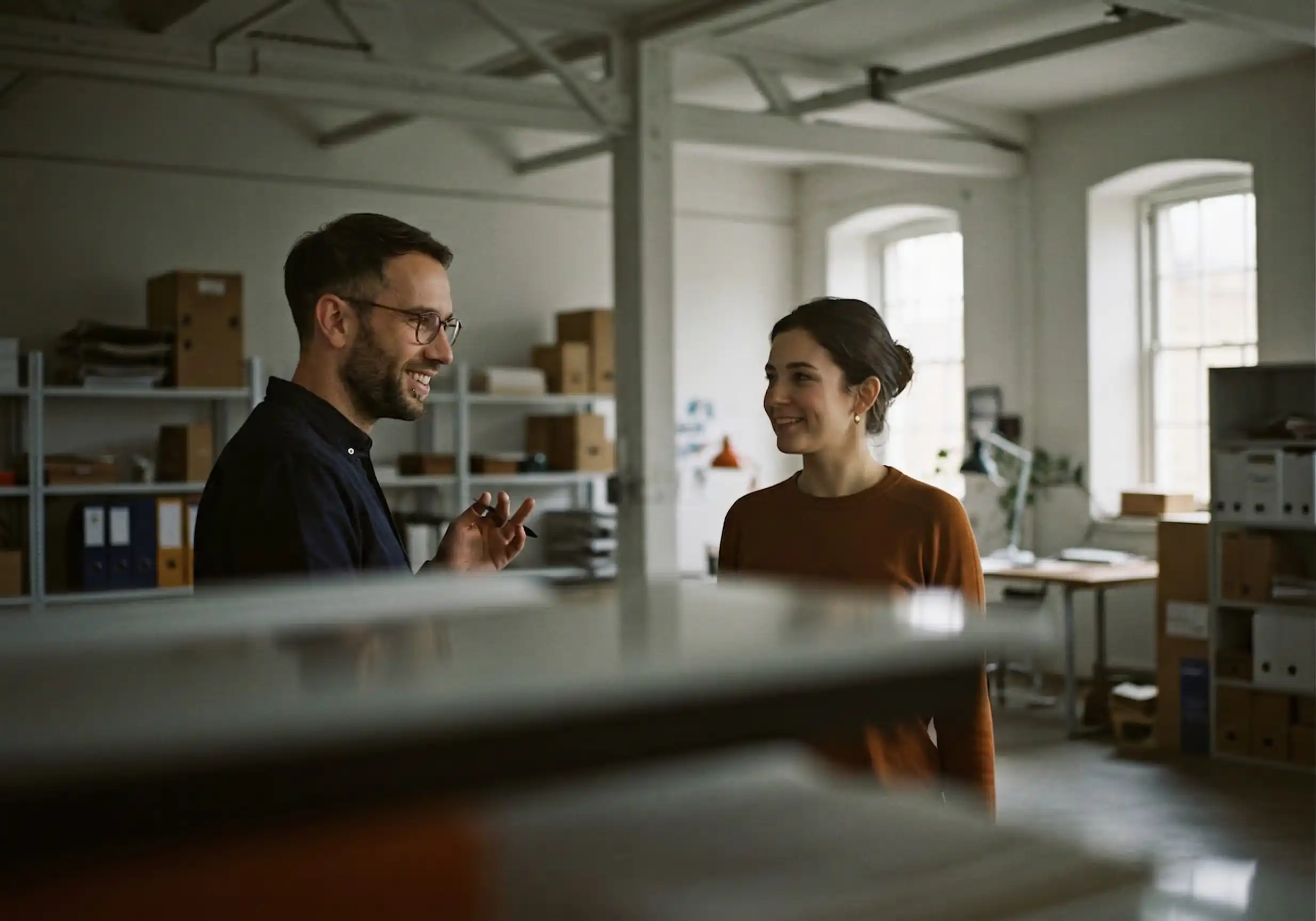 Zwei Kollegen unterhalten sich lächelnd in einem hellen Büro mit Regalen und Ordnern.