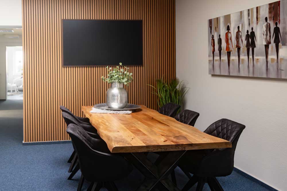 Moderner Besprechungsraum mit Holztisch, sechs schwarzen Stühlen, Pflanzen und großem Bildschirm.
