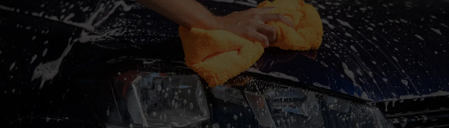 Hand wiping soapy water off a dark car's headlight with a bright orange cloth.