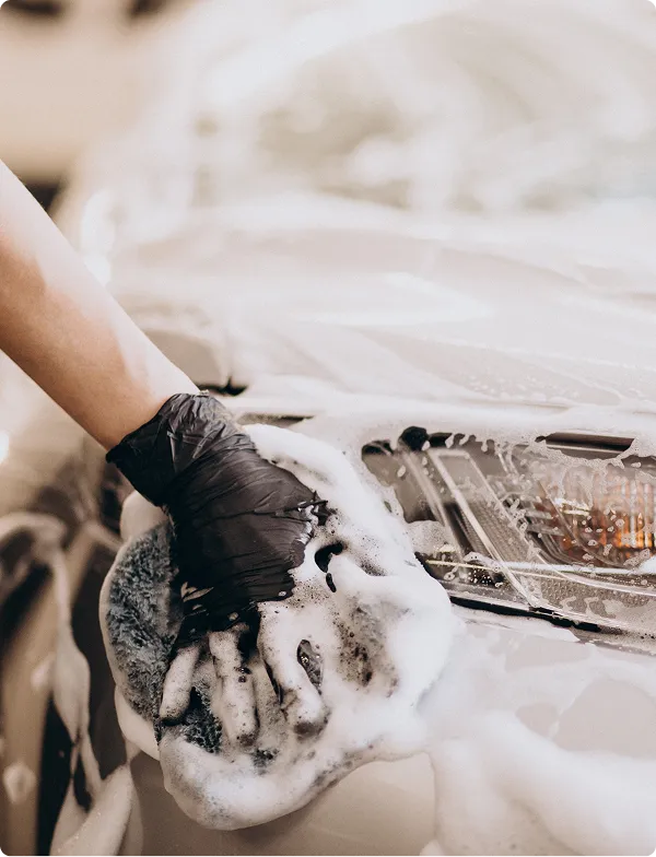 Person wearing a black glove washing a white car with a soapy sponge near the headlight.