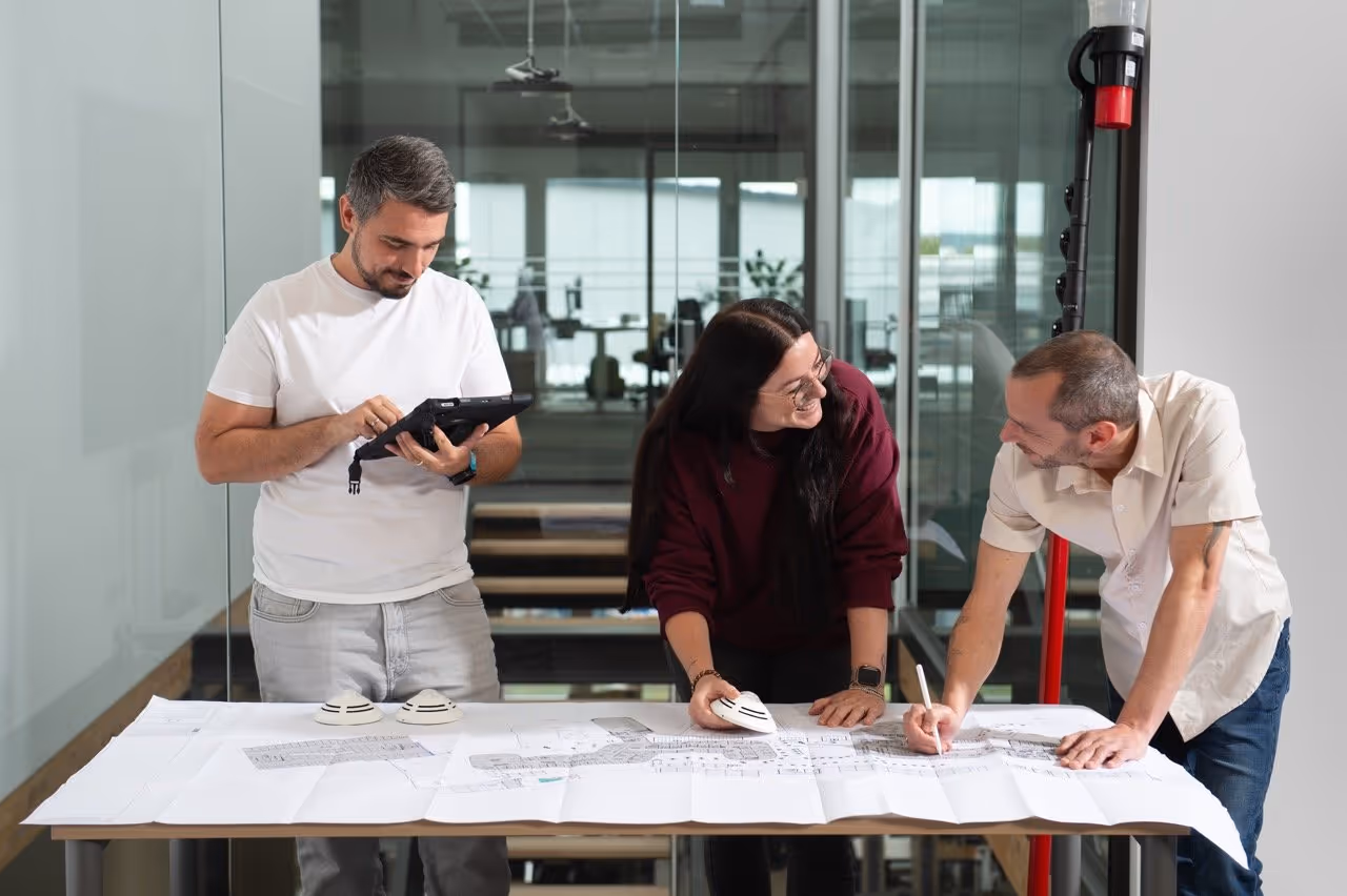 Three Febsfire colleagues discussing architectural plans on a large table, one using a tablet, one holding a model, and another writing on the plans.