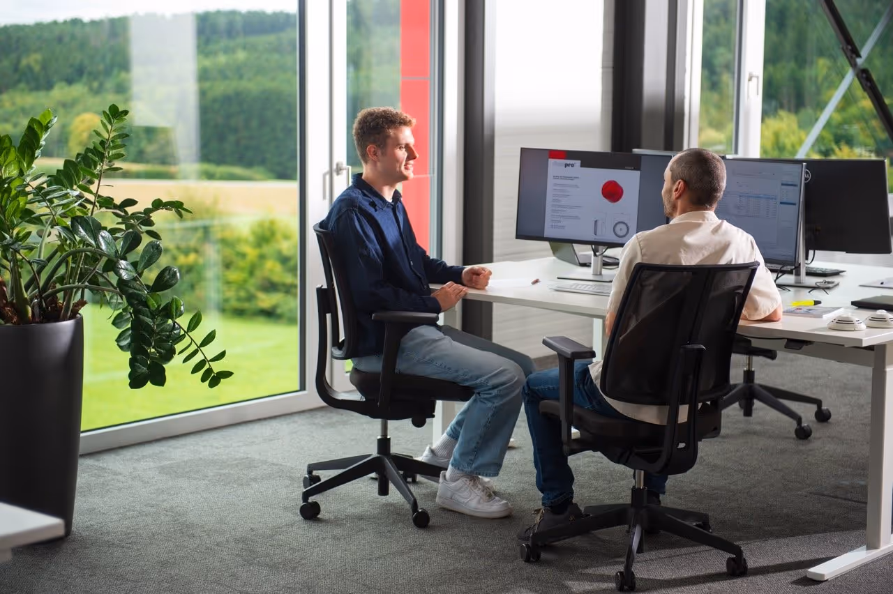 Two men from Febsfire sitting in office chairs having a discussion at a desk with computer monitors, near large windows overlooking a green landscape.