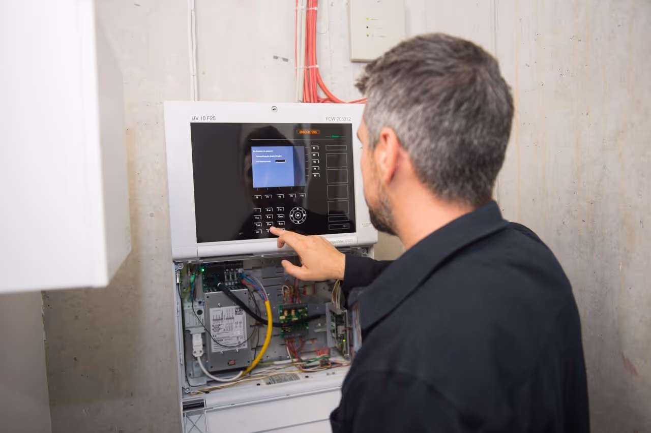Febsfire Technician in black jacket operating a control panel with keypad and electronic components inside an open electrical cabinet.