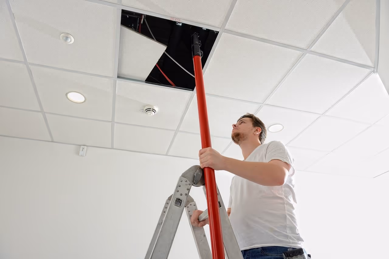 Febsfire technician on a ladder using a red pole to work on a removed ceiling tile in a white-tiled ceiling.