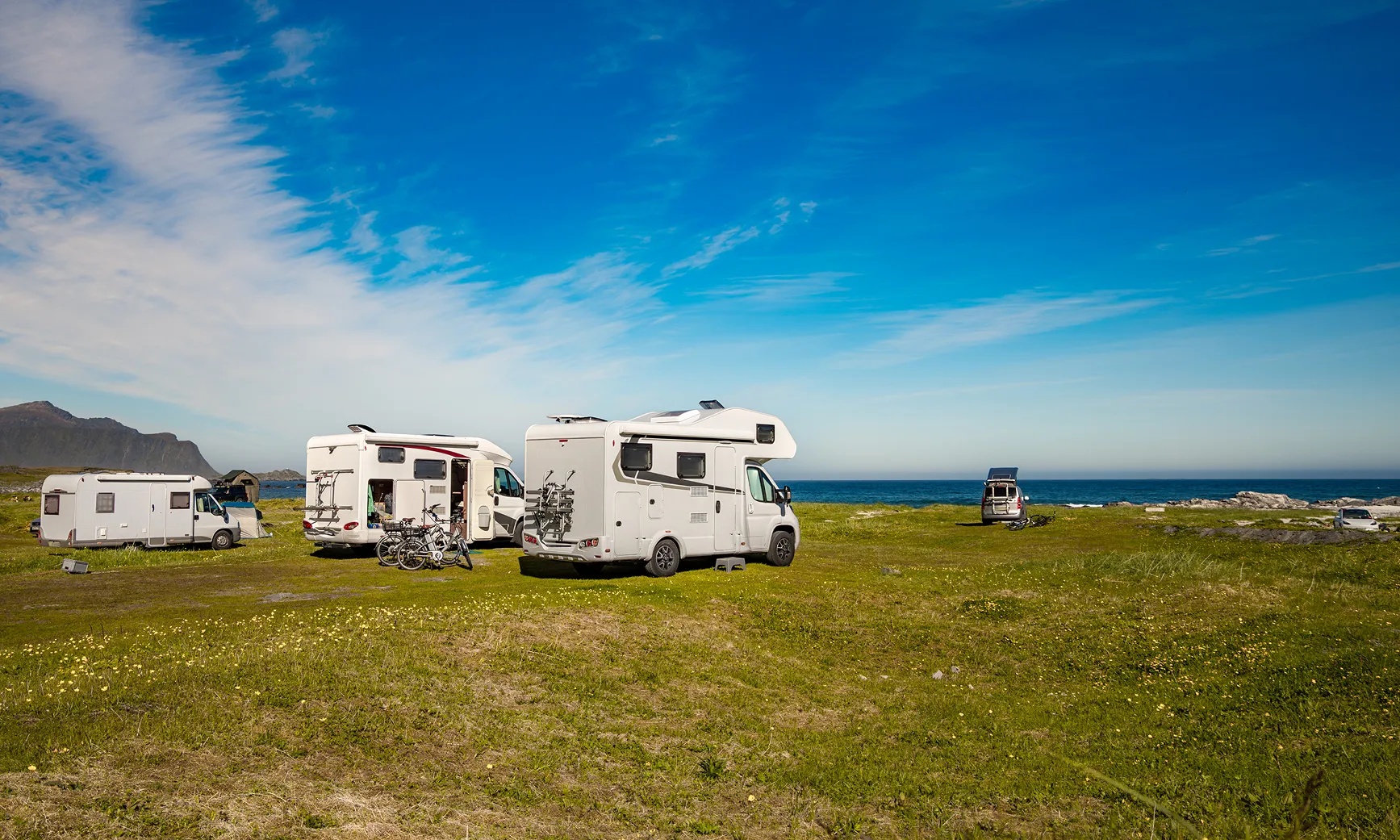 Wohnmobile und Vans auf einer grünen Wiese mit Blick auf das Meer und bewölkten blauem Himmel.