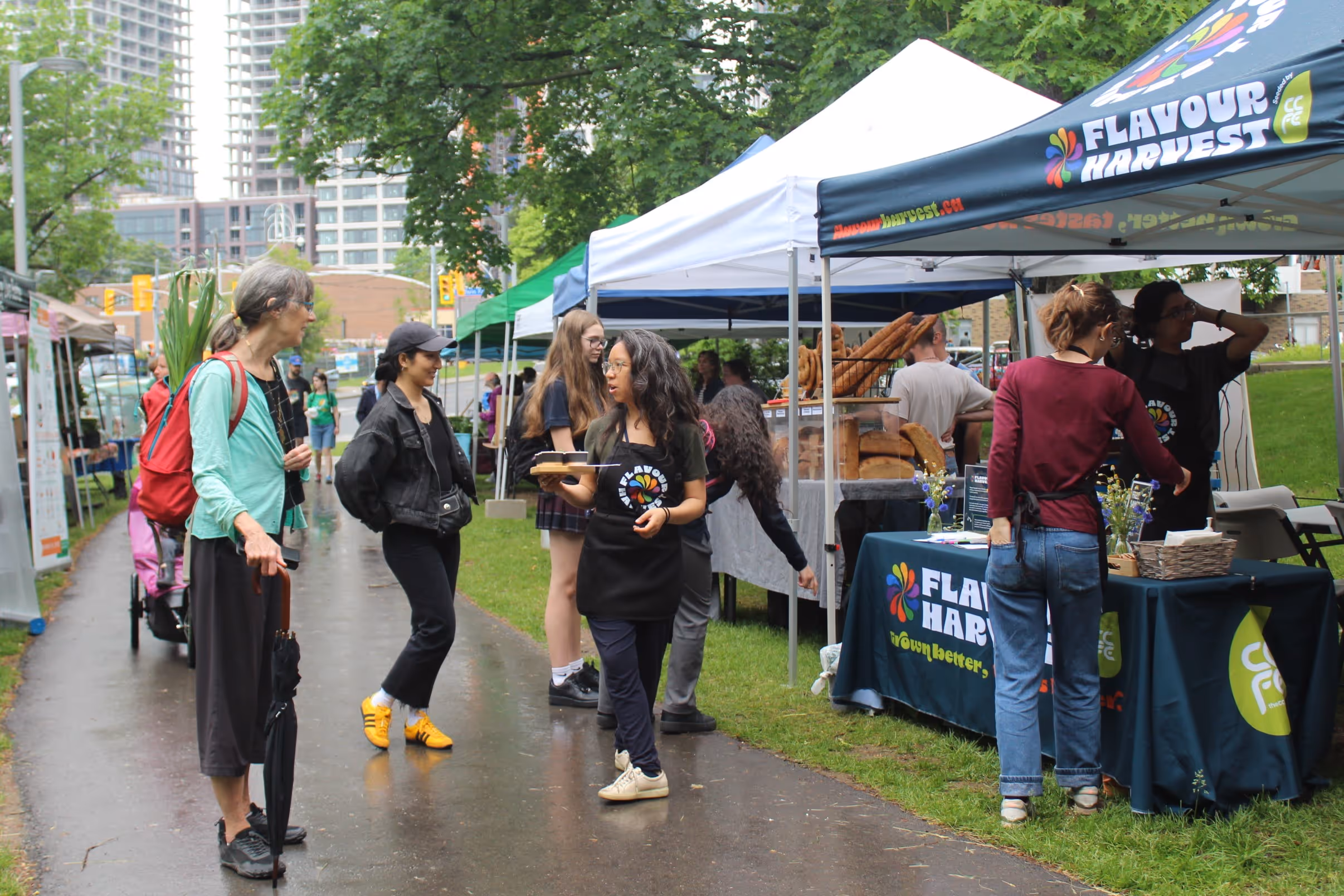 People browsing and serving food at an outdoor Flavour Harvest market stall on a rainy day.