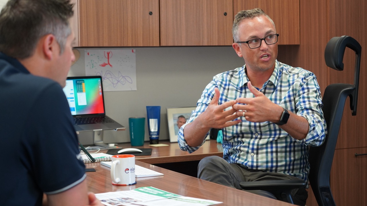Two men having a discussion in an office, one gesturing with hands while seated at a desk with a laptop and documents.