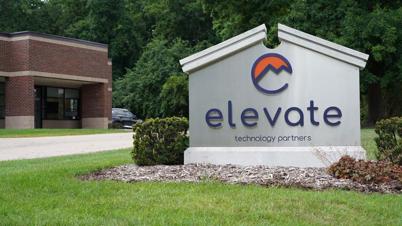 Outdoor sign with logo and text reading 'elevate technology partners' in front of office building and green trees.