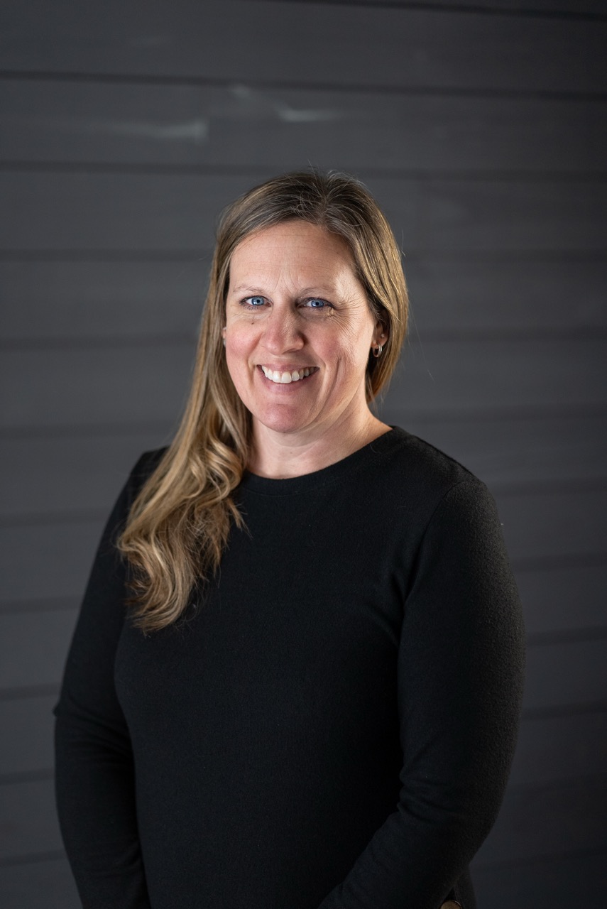 Smiling woman with long blonde hair wearing a black sweater against a dark gray wooden background.