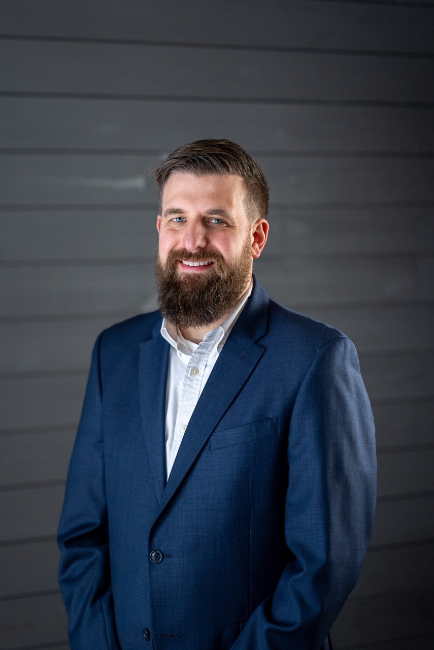 Smiling man with a beard wearing a blue suit jacket and a white shirt standing against a dark gray wooden wall.