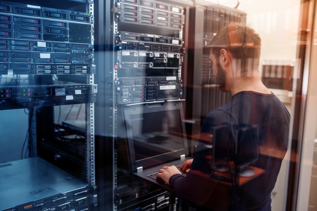 Man working on a laptop inside a server room surrounded by racks of servers.
