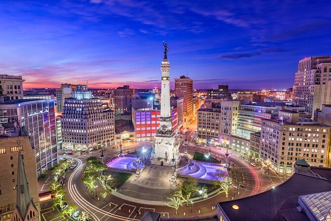 Aerial view of Indianapolis Circle with the Soldiers and Sailors Monument illuminated at dusk, surrounded by city buildings and light trails from traffic.