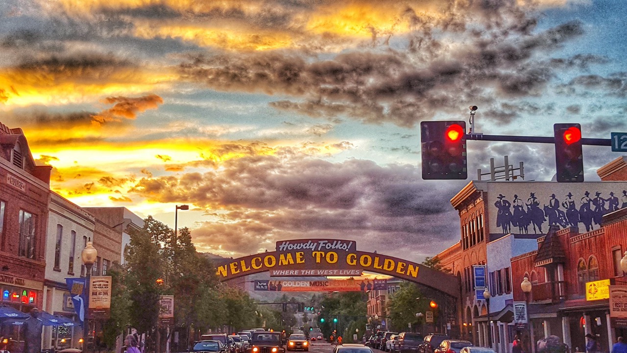 Sunset over a busy street in Golden, Colorado, with a large archway reading 'Welcome to Golden' and traffic signals showing red lights.