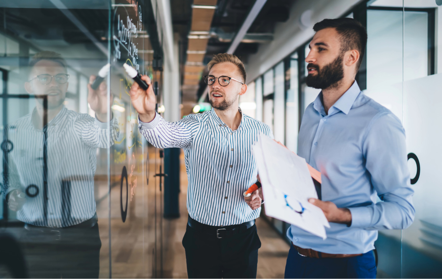 Two men in an office collaborating with one writing on a glass wall while the other holds documents.