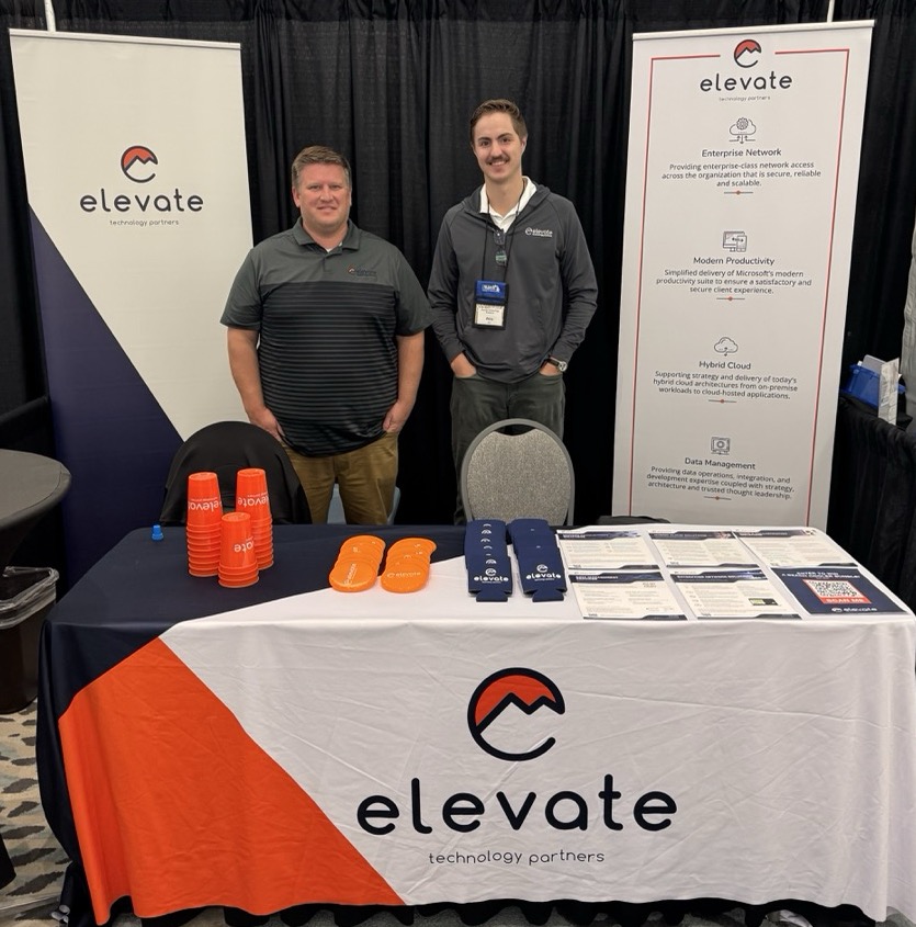 Two men standing behind a trade show table with Elevate Technology Partners branding, including banners, promotional cups, and printed materials.