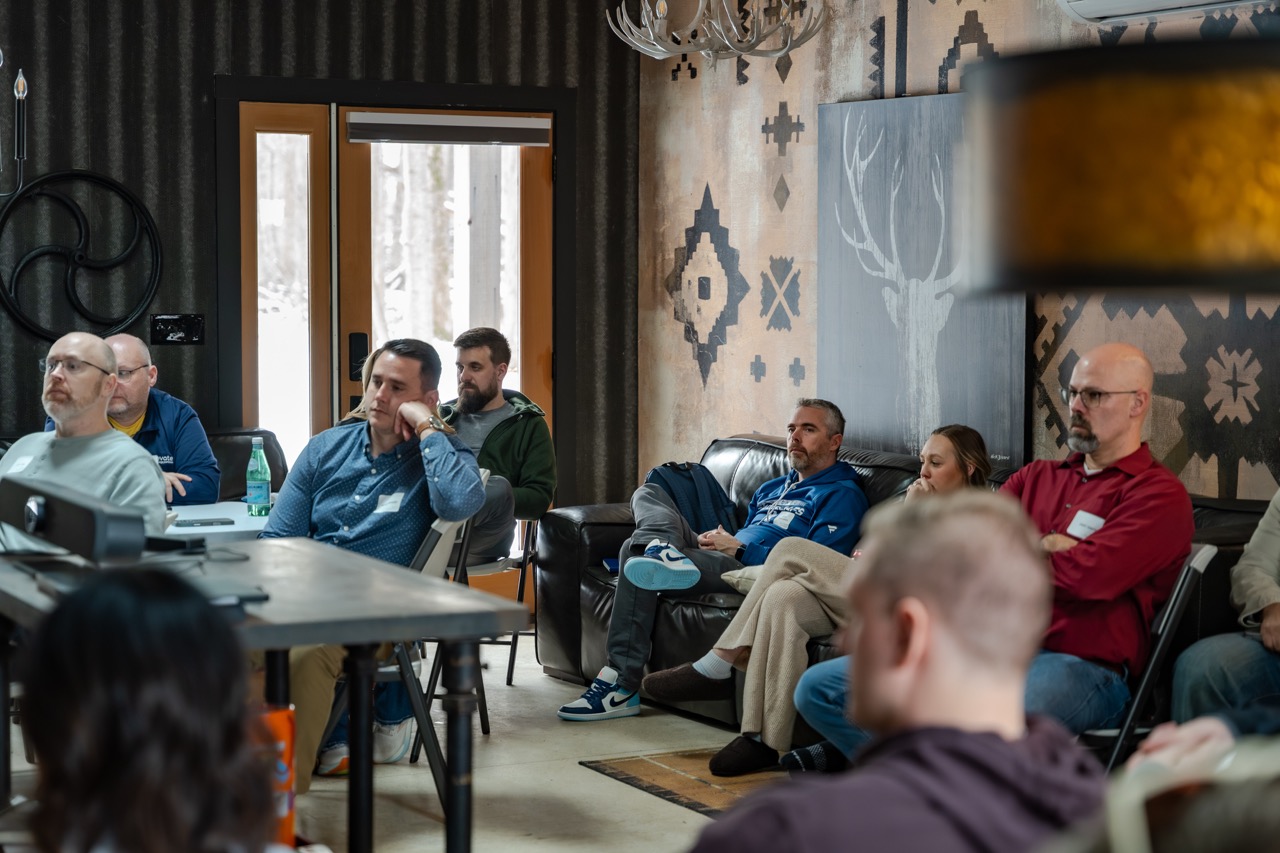 A group of adults attentively listening in a cozy room with patterned walls and wooden doors.