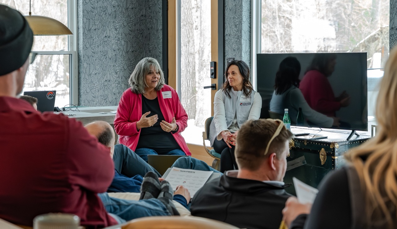 Group of people seated indoors in a casual meeting, with two women speaking near a door and others listening attentively.