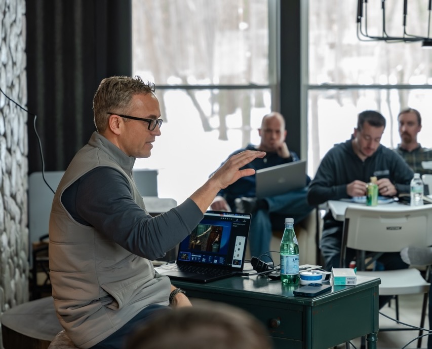 Man in glasses and a vest gestures while seated at a table with a laptop in a room where three other men work on laptops.