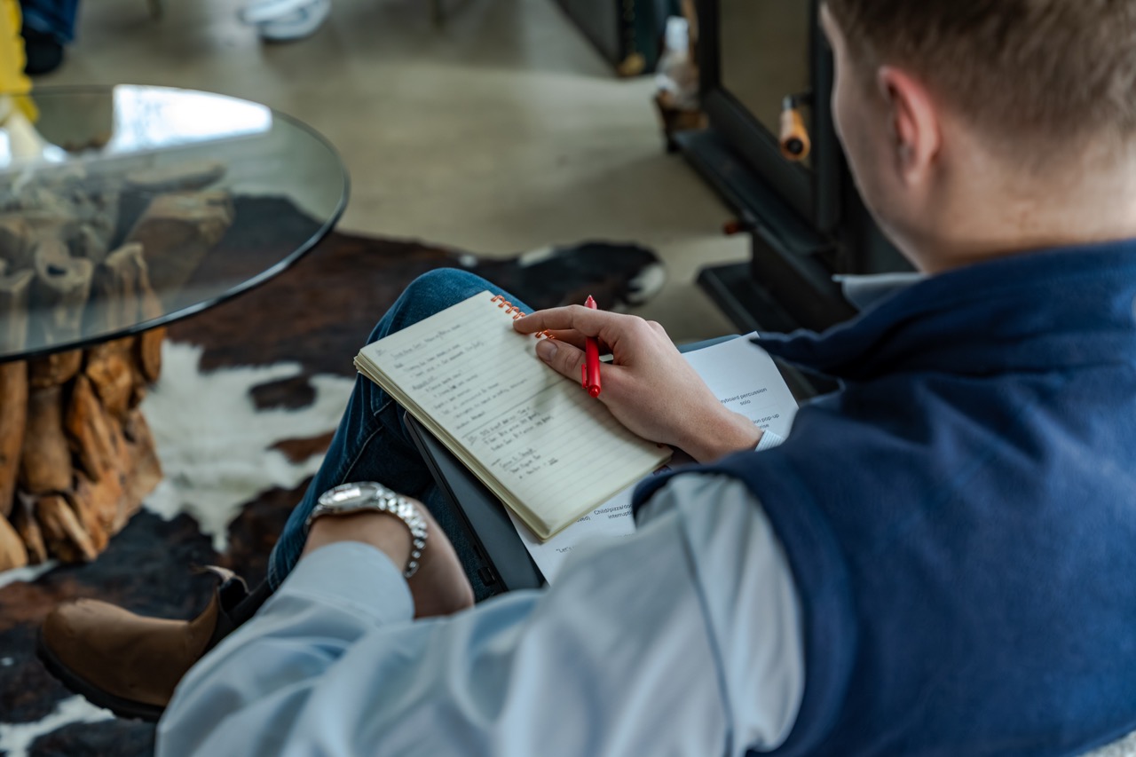 Person sitting indoors holding a red pen and a notebook with handwritten notes, next to a glass table and a cowhide rug.