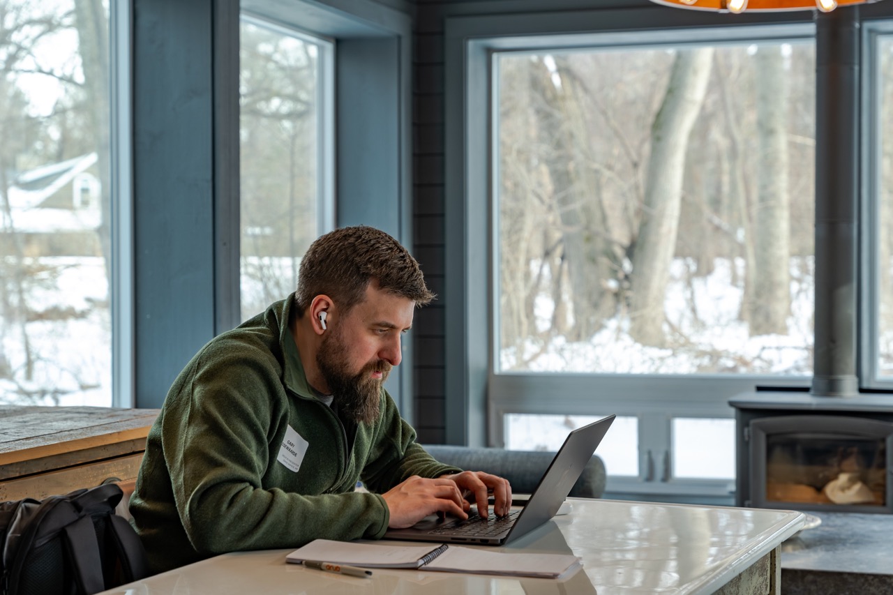 Man in green jacket typing on a laptop at a white table inside a room with large windows showing a snowy landscape.