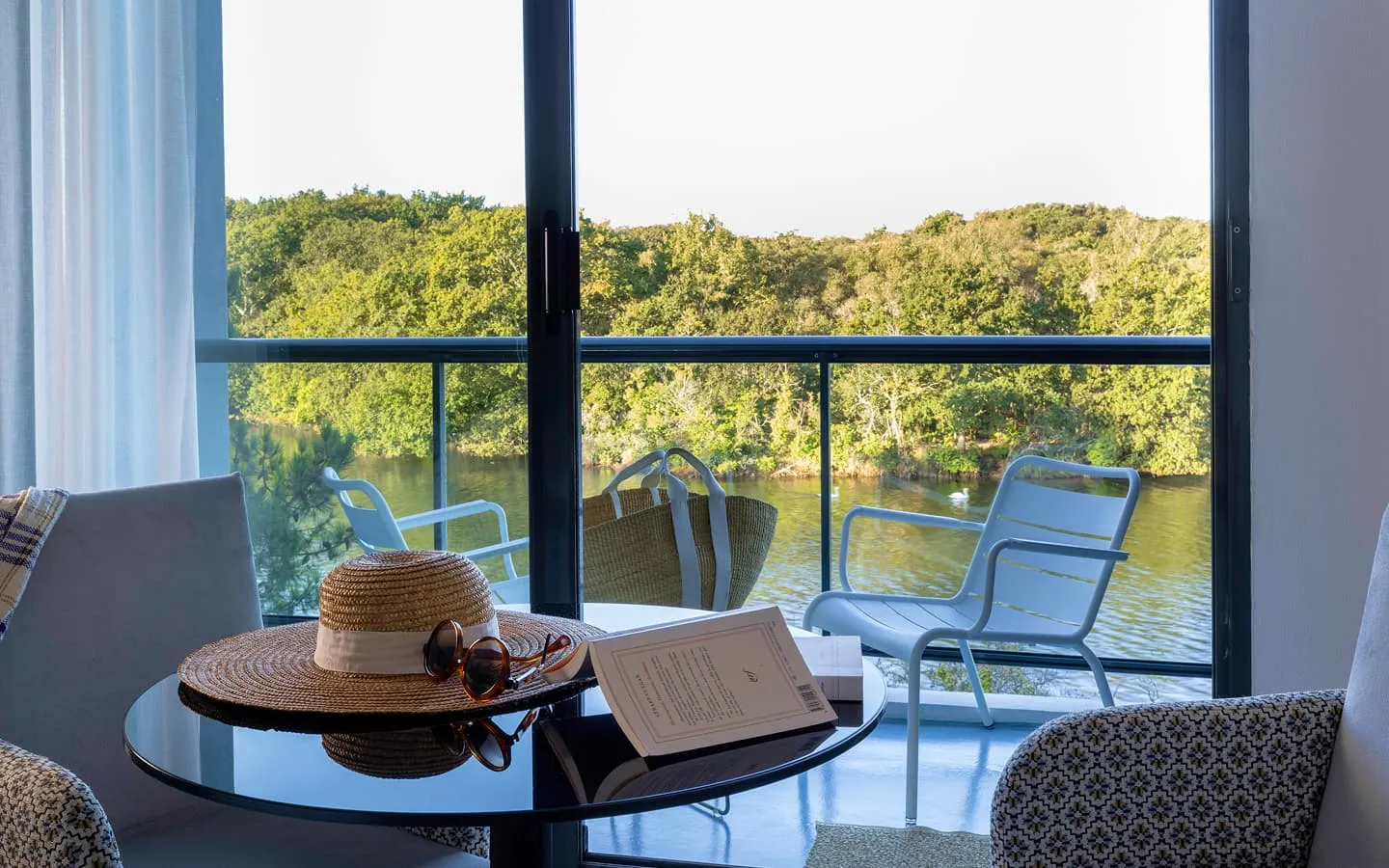 View of the lake surrounded by trees from a terrace with a table, a straw hat, sunglasses, an open book, and two armchairs.