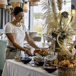 Waitress at La Terrasse du Ter restaurant setting a table with pastries and floral decorations in a bright space.