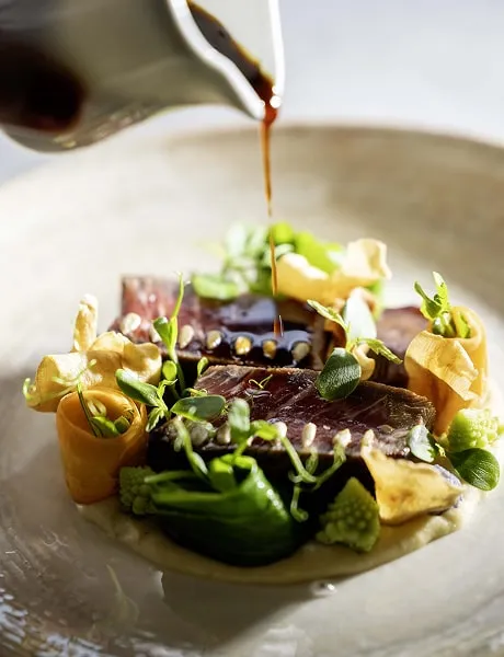 Gourmet dish from La Terrasse du Ter restaurant featuring a slice of meat topped with a dark sauce, accompanied by green vegetables and decorated with edible leaves and flowers.