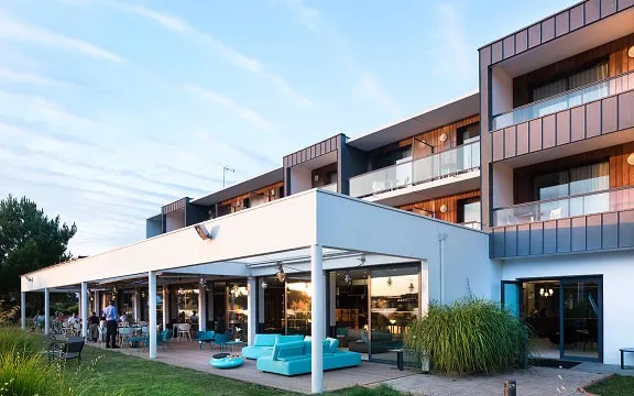Modern façade of the Rives du Ter hotel with outdoor terrace furnished with turquoise blue sofas, under a clear sky at sunset.
