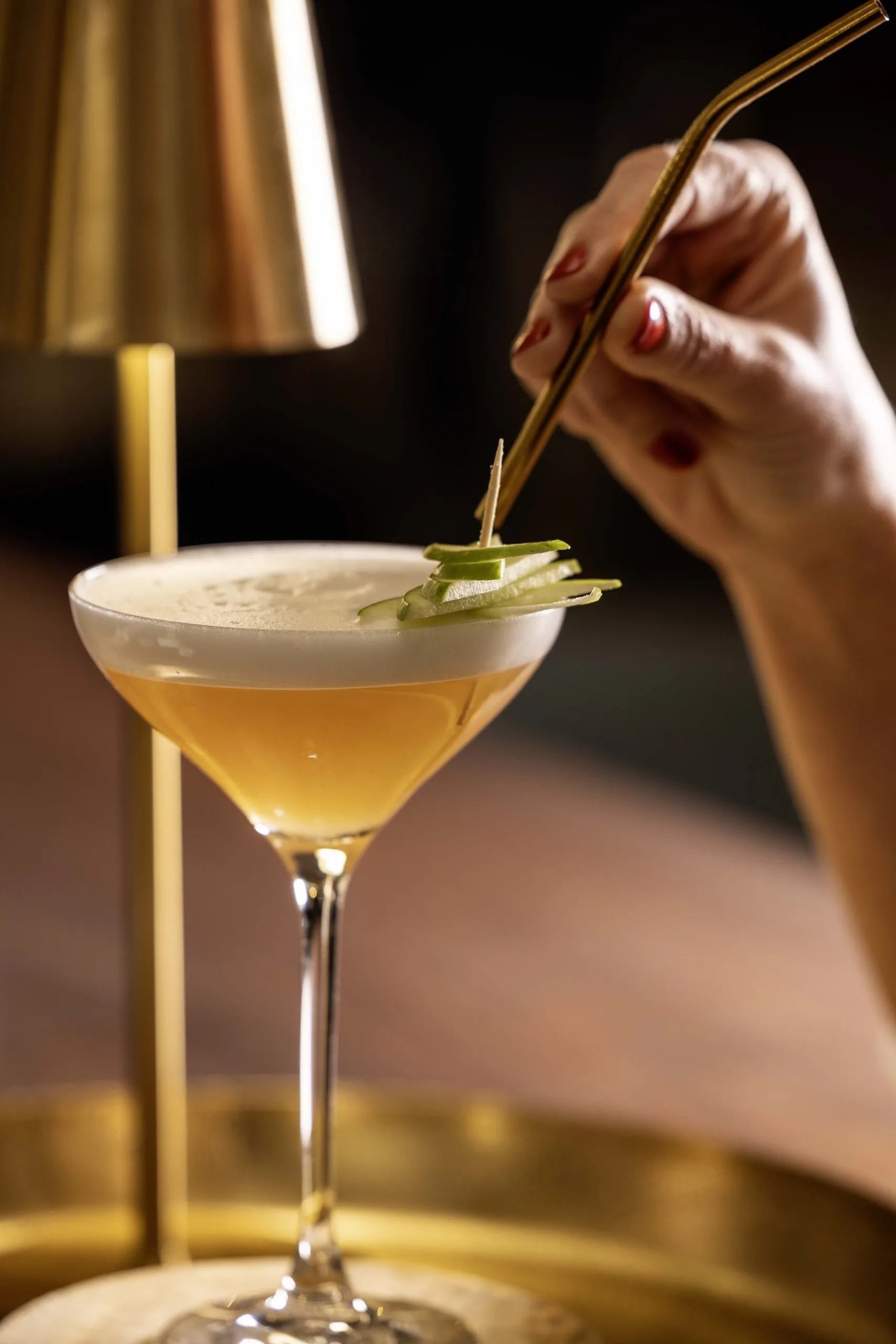 Golden cocktail with white foam, decorated with thin slices of green apple on a toothpick, held by a hand with red nail polish, on a golden tray.