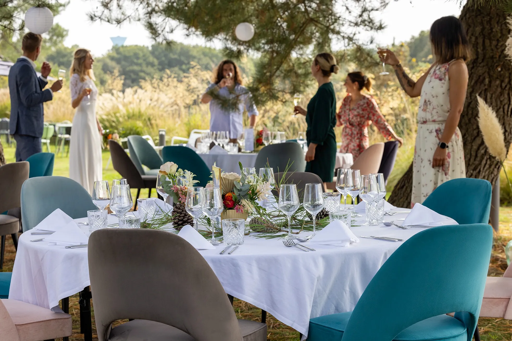 Guests celebrating a wedding outdoors around a reception table with white tablecloths and floral decorations under a large tree.