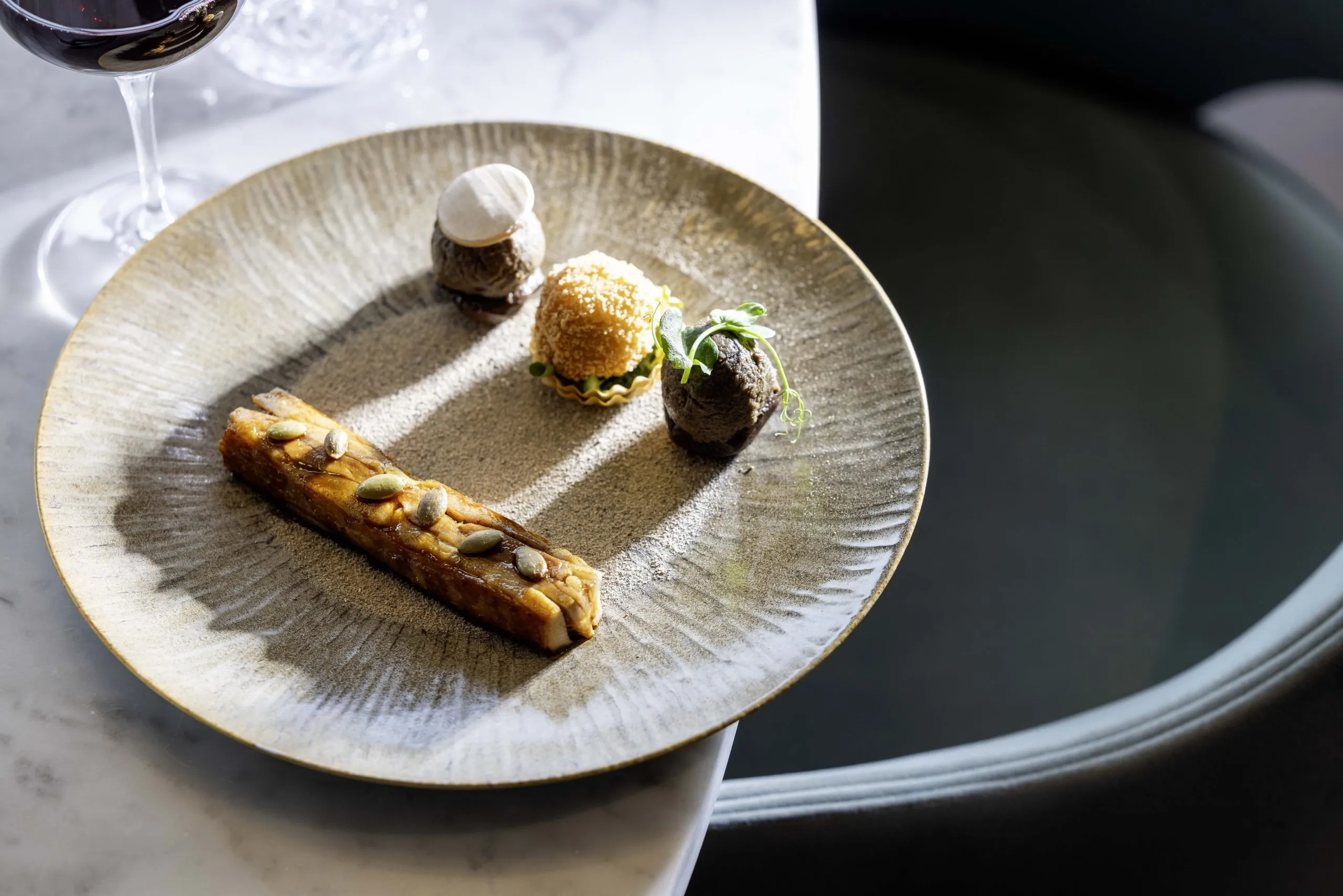 Gray plate with a refined presentation consisting of a golden rectangle garnished with seeds, three small artistic bites, and a glass of red wine on the left.