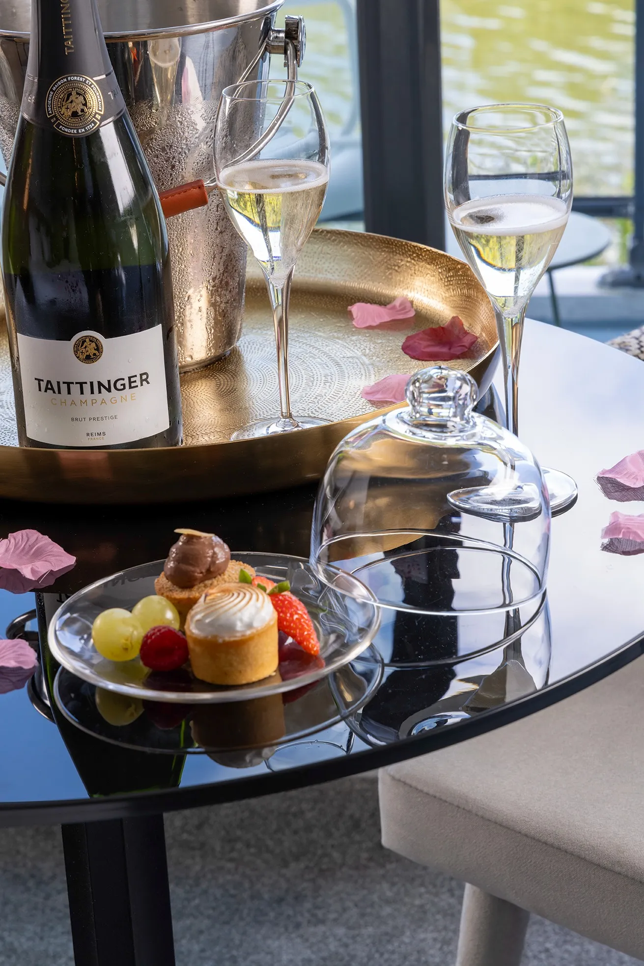 Bottle of Taittinger champagne with two filled flutes, desserts, and fruit on a glass plate on a black table with rose petals.