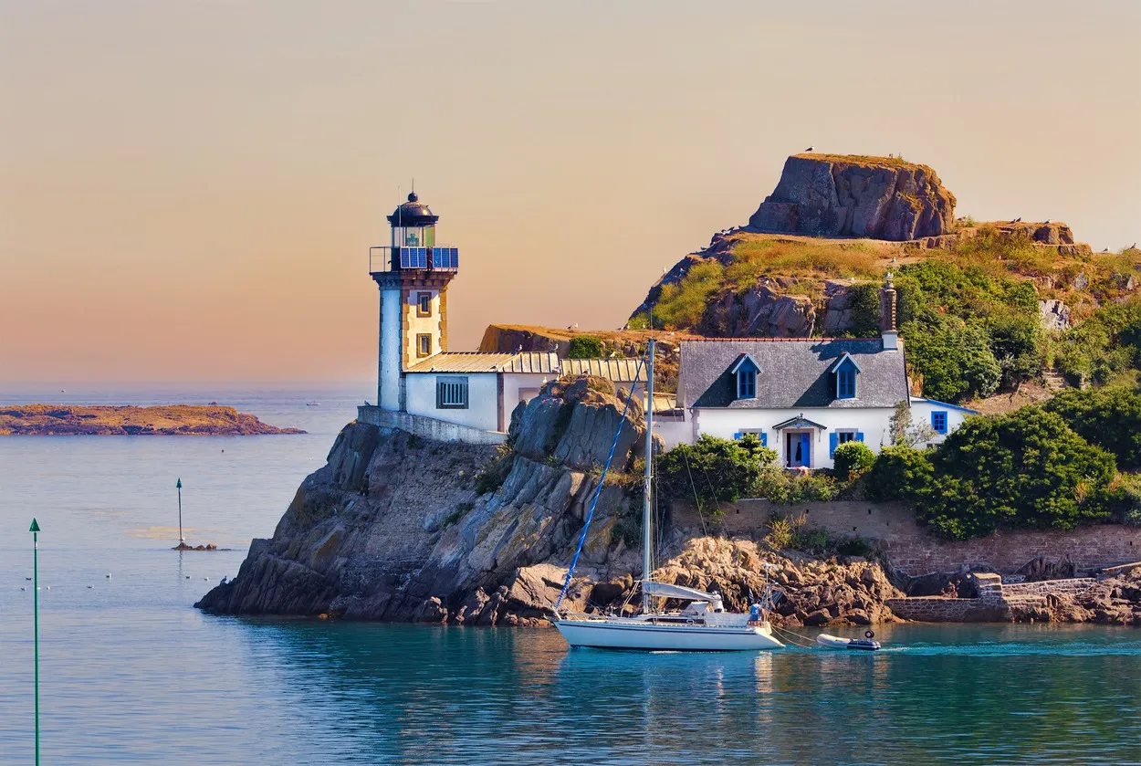 White lighthouse and house near a rocky cliff by the water with a sailboat anchored nearby at sunset.