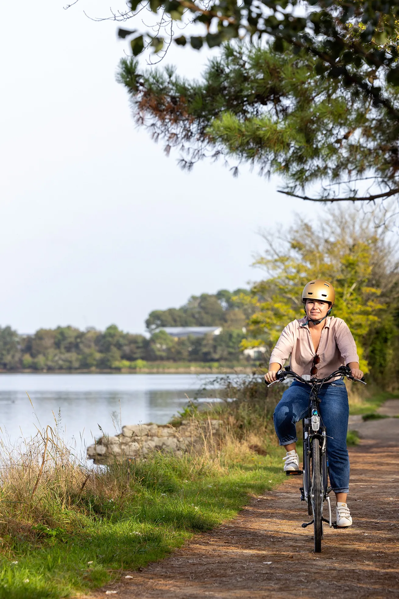 Woman wearing a gold helmet riding a bike on a lakeside trail, surrounded by trees and vegetation.
