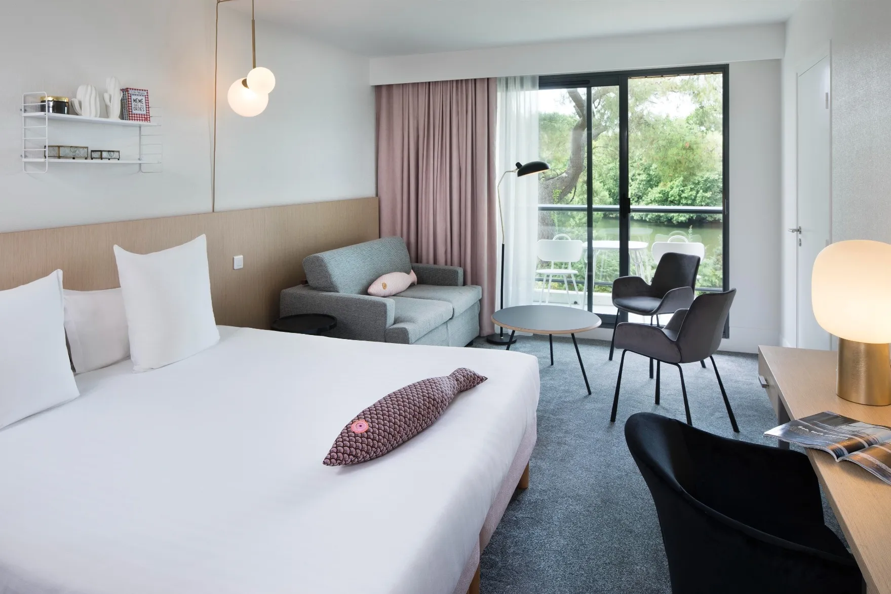Room at the Rives du Ter hotel with king-size bed, gray sofa, two armchairs, coffee table, desk with lamp, and view of a balcony with white chairs and trees.