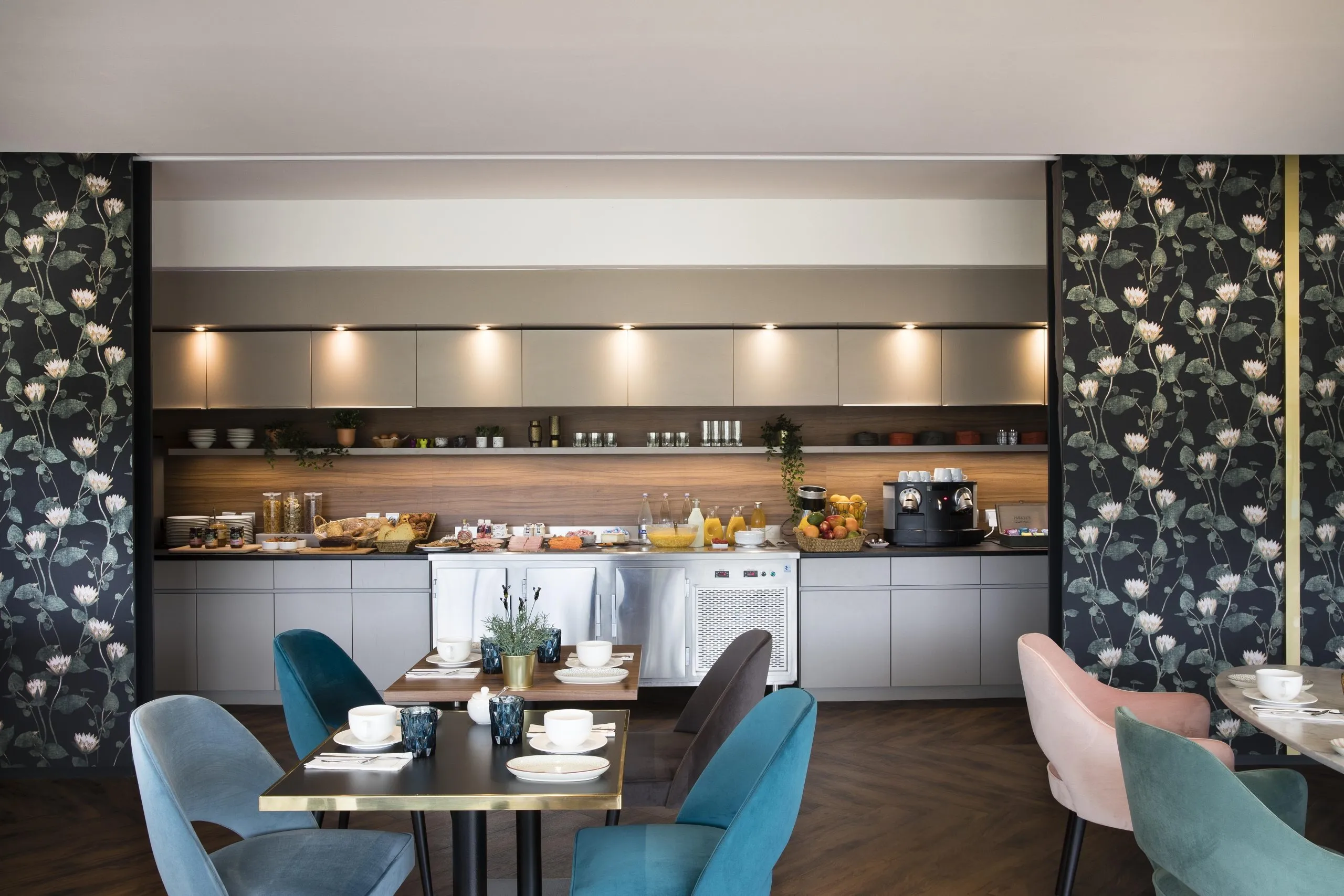 Modern breakfast area at the Rives du Ter hotel with a buffet including juice, fruit, coffee, bread, cereal, and tables with colorful velvet chairs.