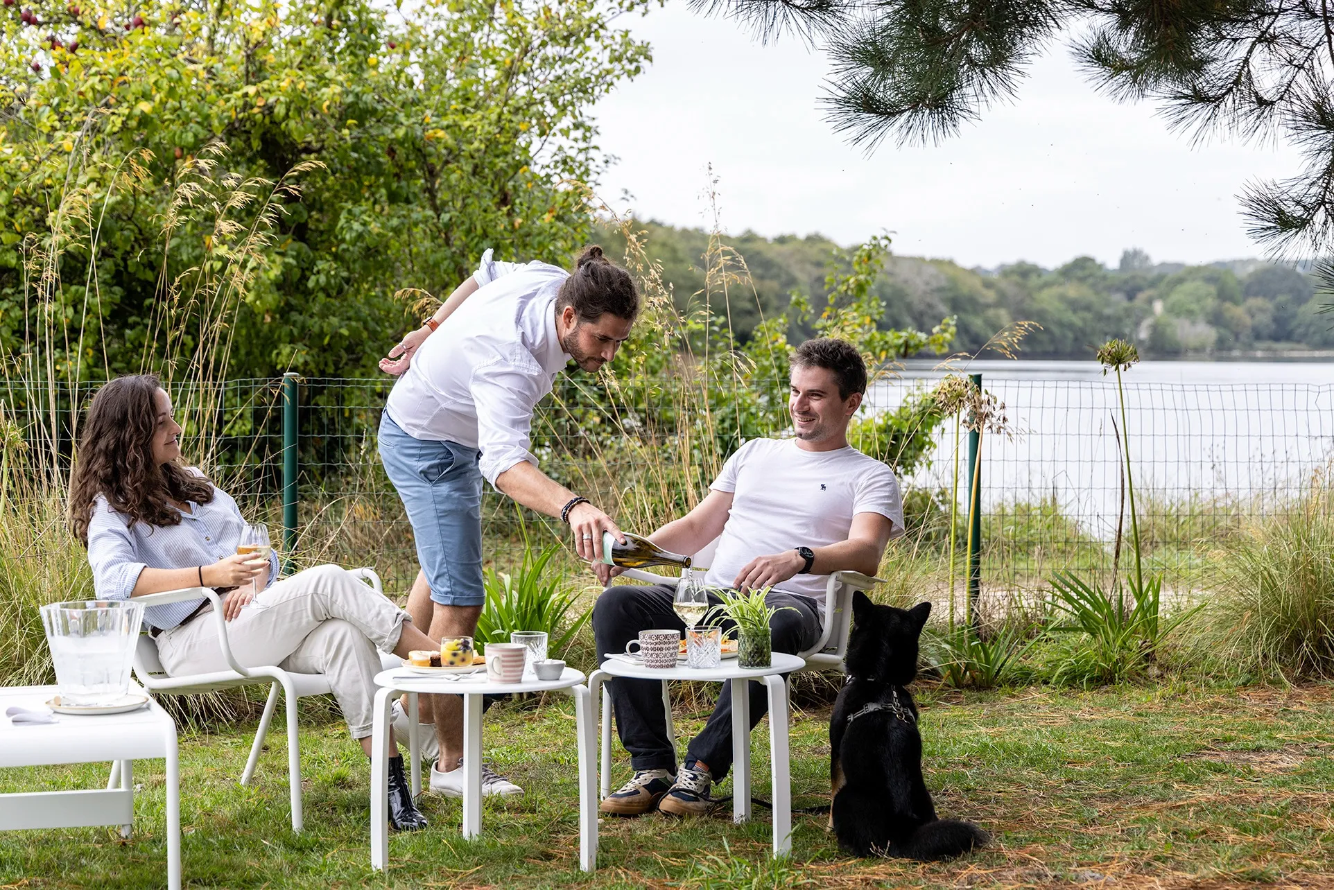 Three people are enjoying some time outdoors near the lake. A man is pouring white wine for another man who is seated, while a woman is holding a glass and a black dog is sitting nearby.
