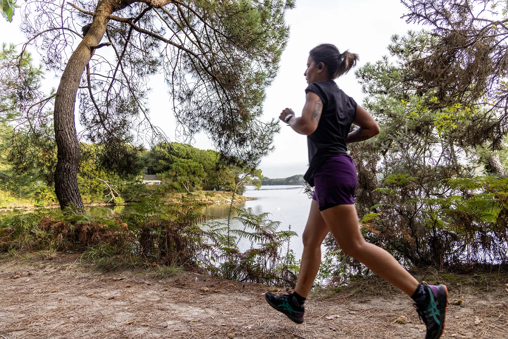 Woman running on a forest trail by the lake surrounded by green trees.