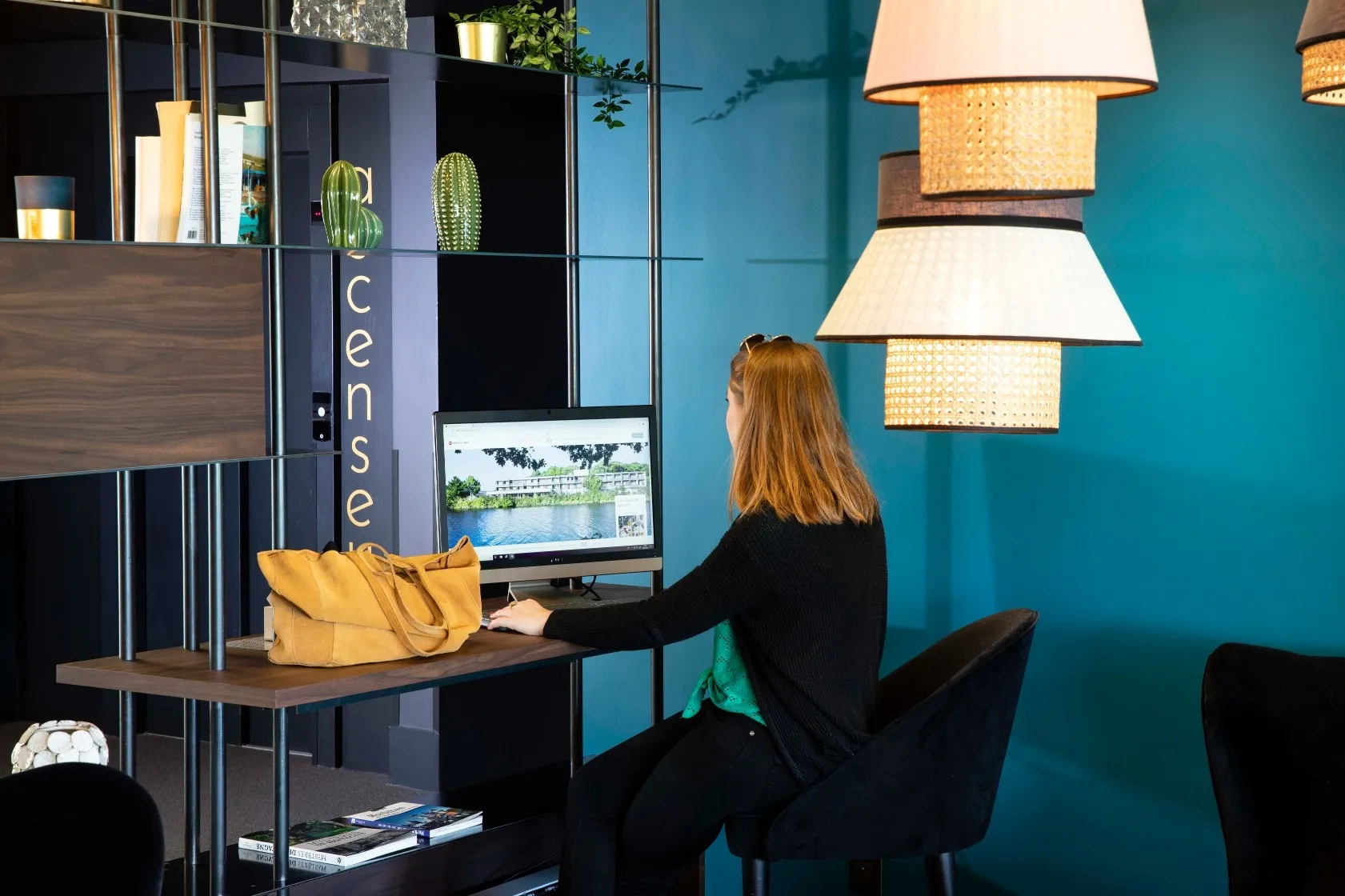 Woman sitting in a black chair working on a computer in a modern space with hanging lights and a yellow handbag on the table.